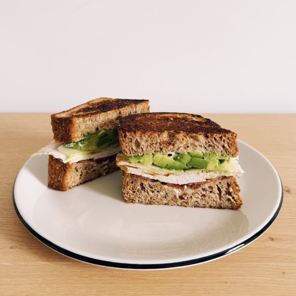 A turkey, avocado and bacon sandwich on toasted seeded bread, halved on a plate.