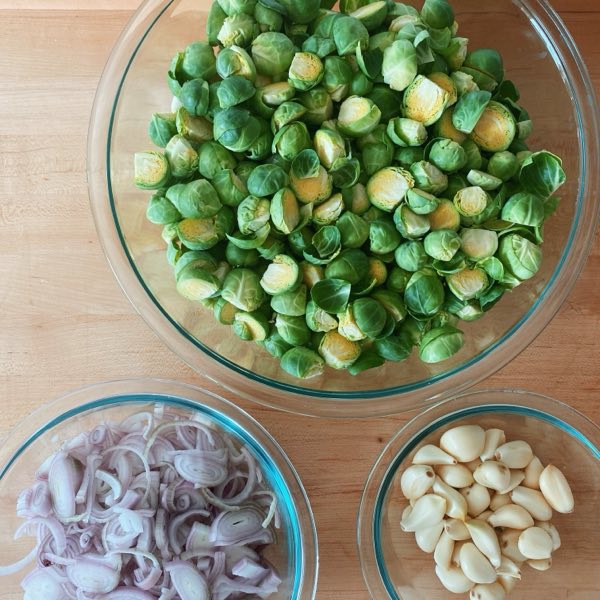 Brussels sprouts, sliced shallots and peeled garlic in glass bowls on a wooden board.